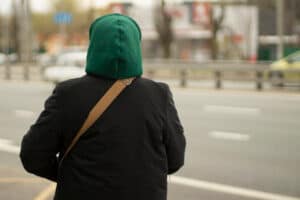 An unknown young person on the road, warmly dressed in a green hood and black jacket. The image is used for PolicyWise for Children & Families' report that research points to childhood roots of youth 