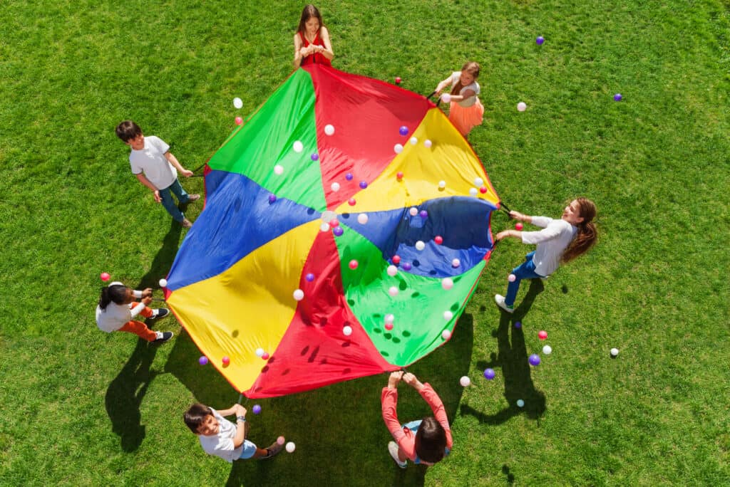 An image for the Open Position - Director of Business & Fund Development - Top view picture of seven kids standing in a circle and waving rainbow parachute in order to throw colorful balls in the air