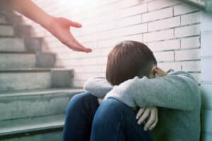 A young man who is homeless sit in an underground stairwell. A hand is reaching to provide them help. This is image is being used for PolicyWise's news story 